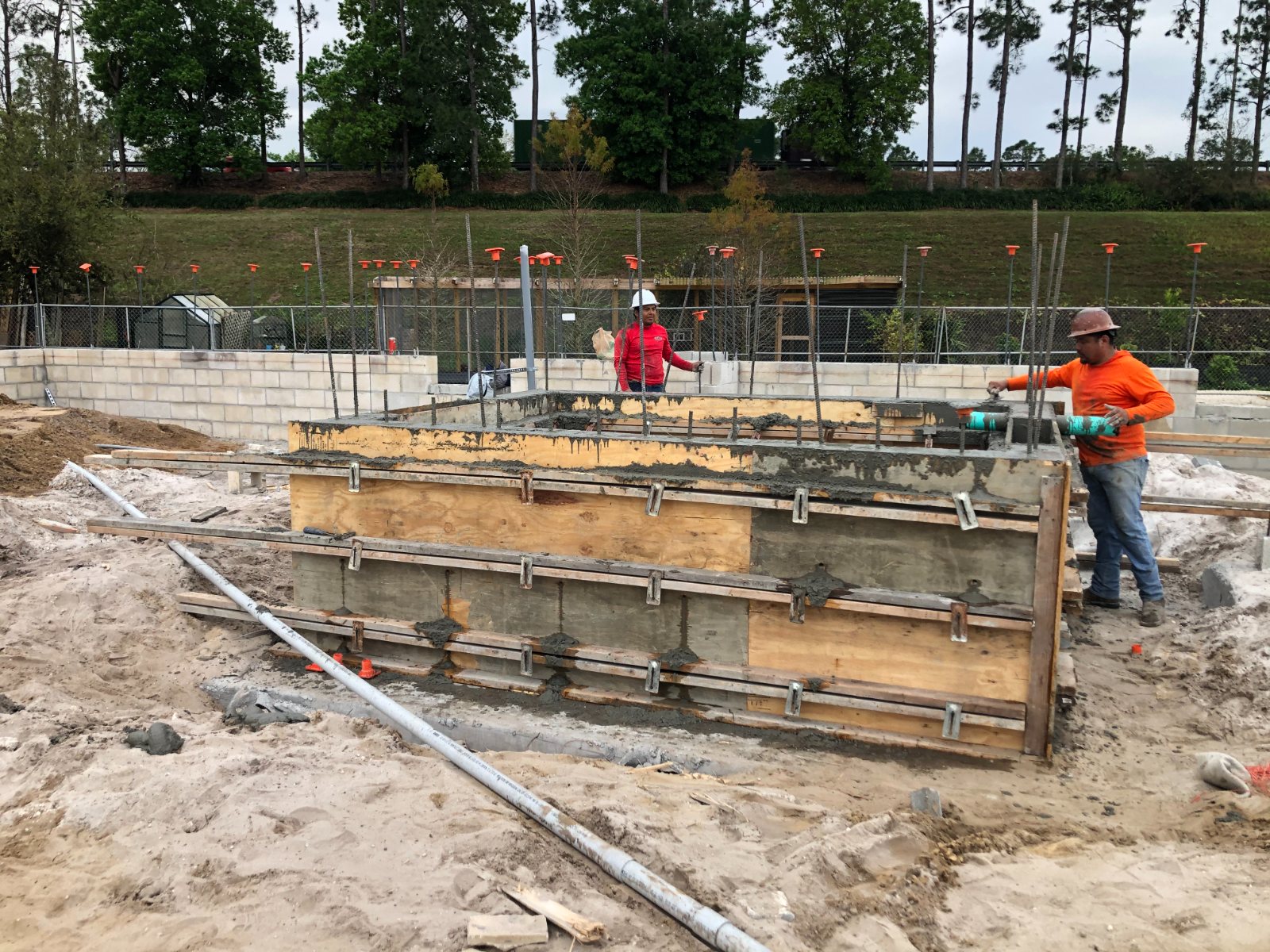 Crew pouring concrete into plywood beam forms, block walls rising behind