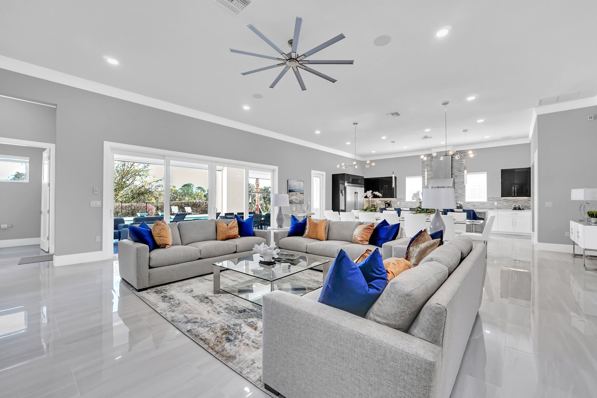 Open great room with grey sectionals, flowing to the chef's kitchen and out to the pool