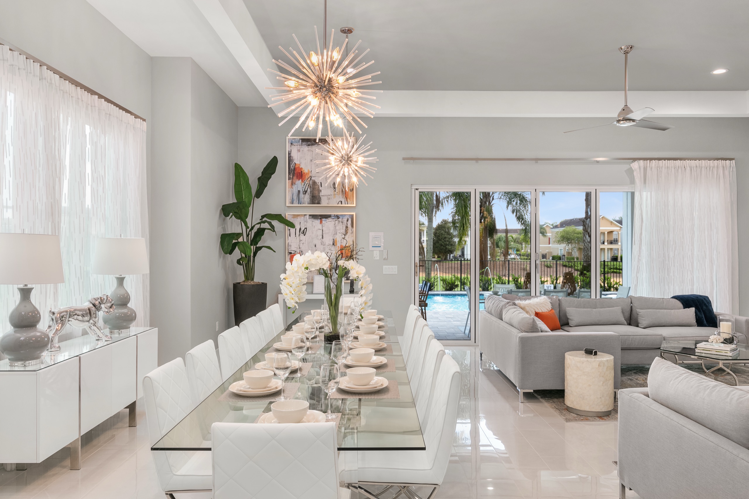 Glass dining table set for twelve under sputnik chandeliers, with the living room and pool beyond